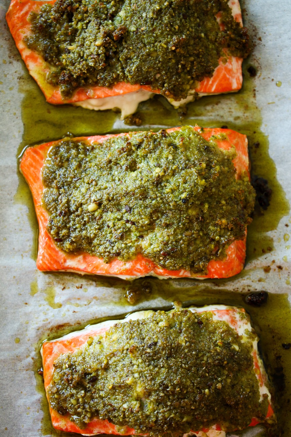 Three bright orange wild salmon fillets with green pesto crust on parchment paper, shot from overhead.