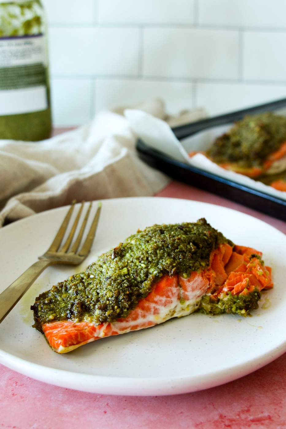Bright orange wild caught salmon, flaked, with a green pesto crust on a white plate, on a pink surface, with a jar of pesto and subway tiles in the background.