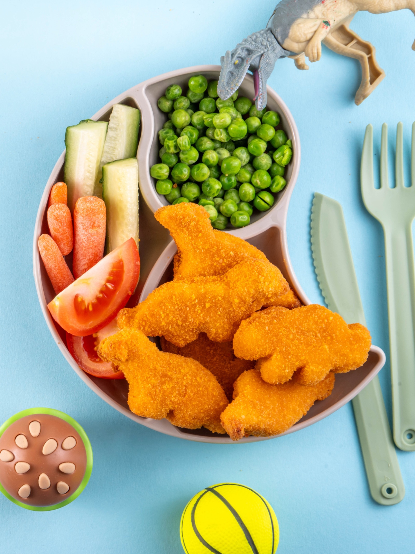 A kid's plate with dinosaur-shaped nuggets alongside peas, cucumber, carrots, and tomato — served on a blue table with toy dinosaurs.