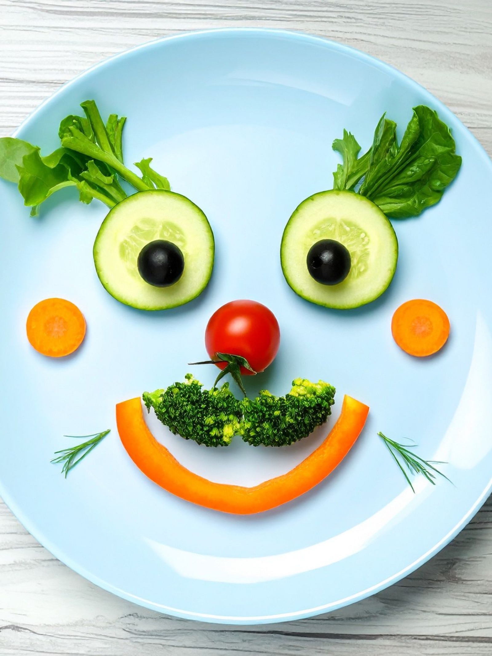 A plate arranged as a smiley face with cucumber eyes, a cherry tomato nose, broccoli mustache, and a bell pepper smile.