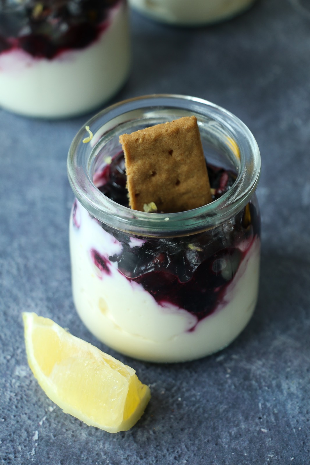 Close-up of a single glass jar filled with creamy lemon cheesecake, mashed blueberries, and a graham cracker garnish, with a lemon wedge on a dark stone surface.