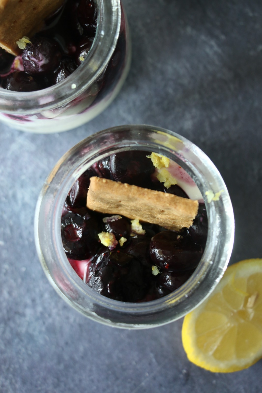 Overhead view of two glass pots filled with creamy lemon cheesecake, mashed blueberries, and a graham cracker garnish, with a lemon slice on the side.