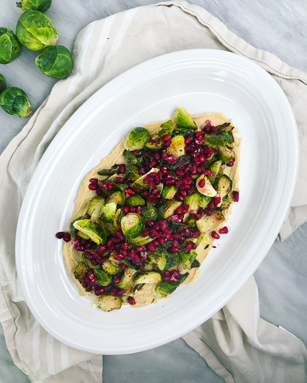 Crispy Brussels sprouts with pomegranate and hummus on a white plate, on a marble background.