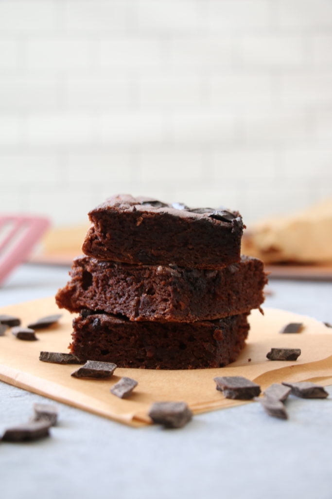 Vertical stack of three chocolate cake bars, surrounded by chocolate chips, on parchment paper with a subway tile backsplash