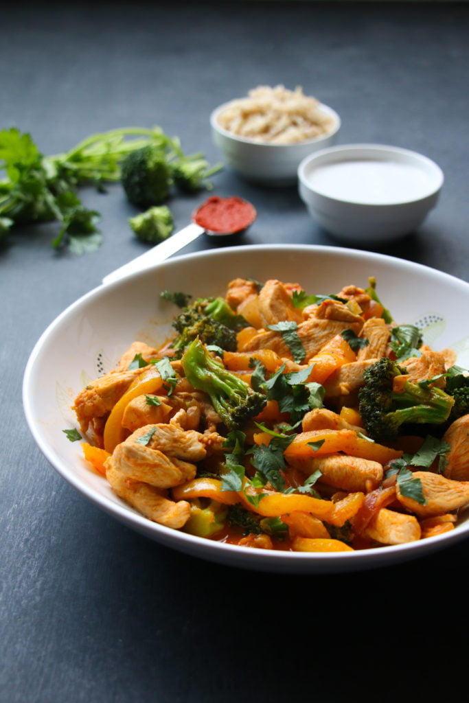 Thai red chicken curry in a white bowl on a black back drop, with rice, coconut milk, broccoli and red curry paste in the background.