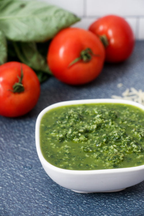 Basil pesto in a white bowl on a blue background, surrounded by basil and tomatoes.