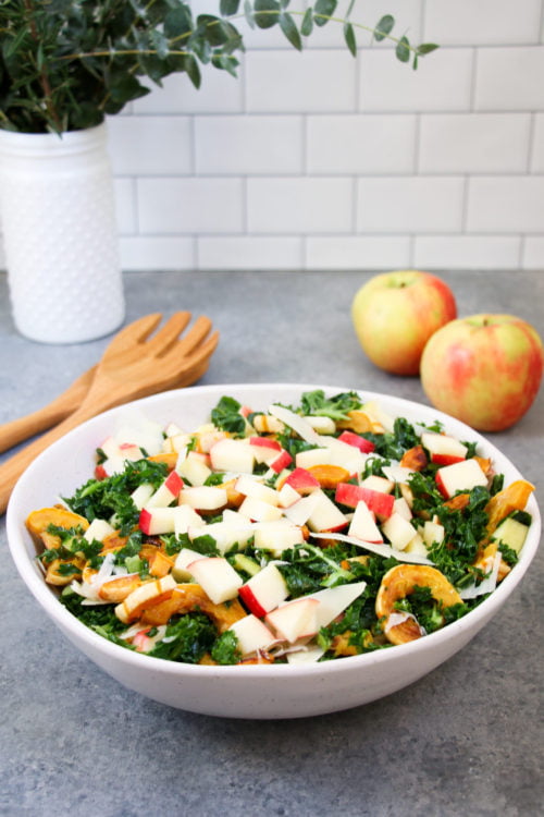 Delicata squash and apple kale salad with honey mustard dressing in a white bowl on a gray background