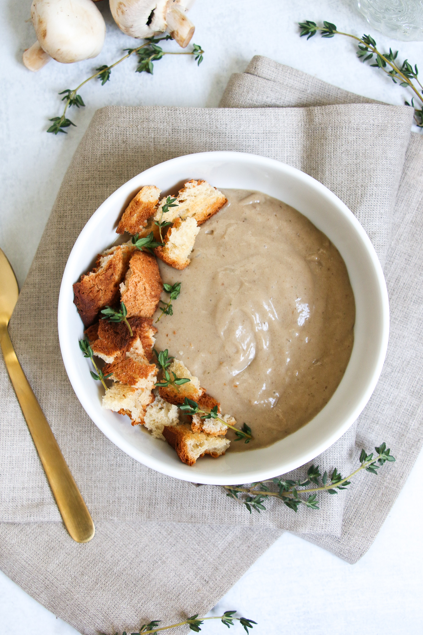 Vegan cream of mushroom soup, topped with croutons and thyme in a white bowl on a gray and white background.