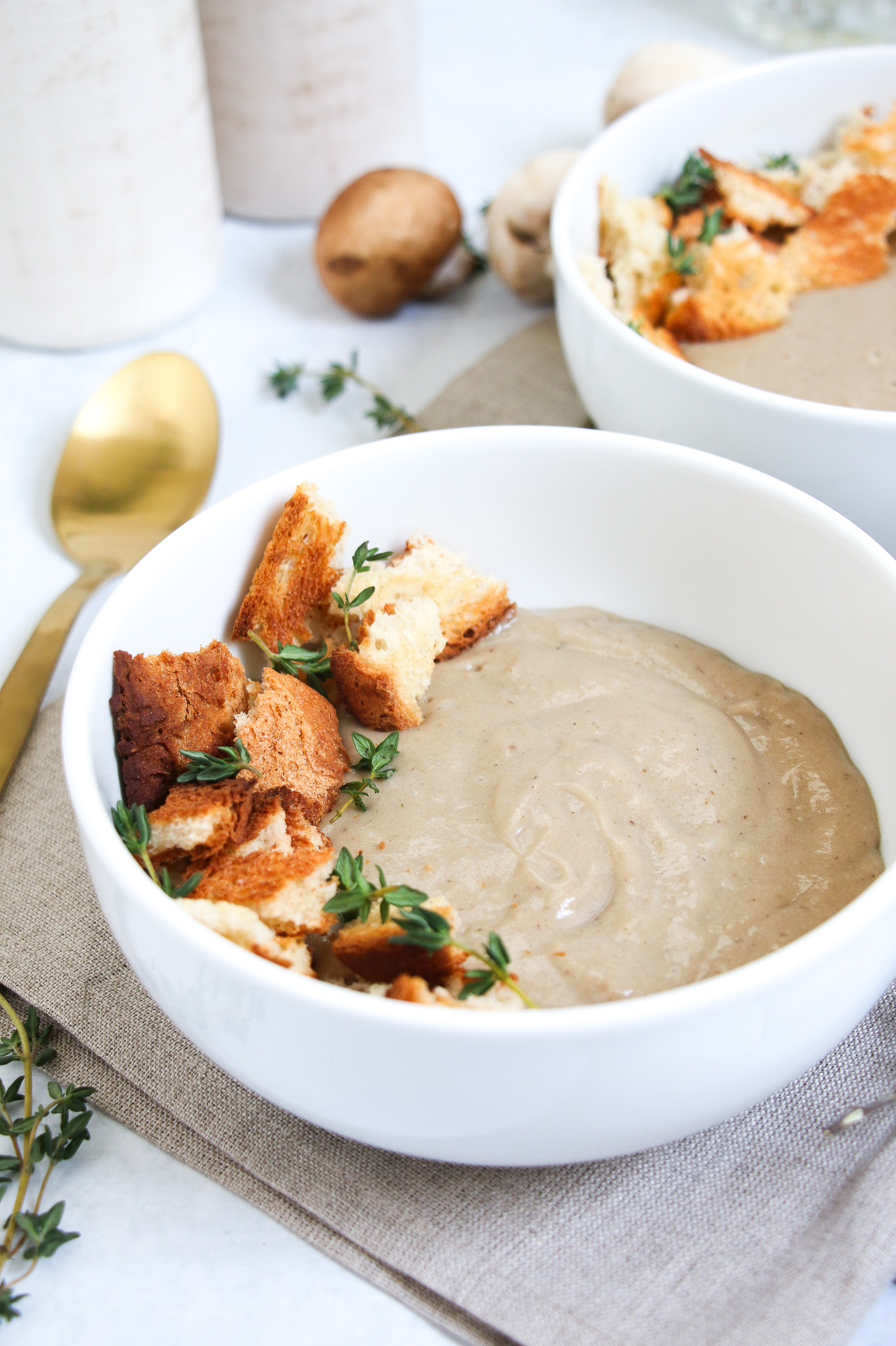2 bowl of healthy, cream of mushroom soup, topped with croutons and thyme in a white bowl.