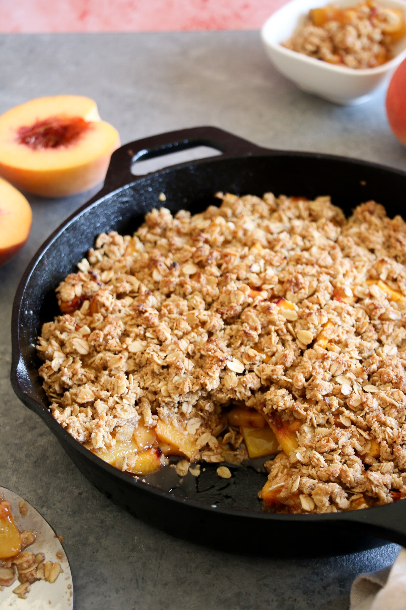 gluten-free peach crisp made with oats in a cast iron pan on gray background with peaches behind the pan